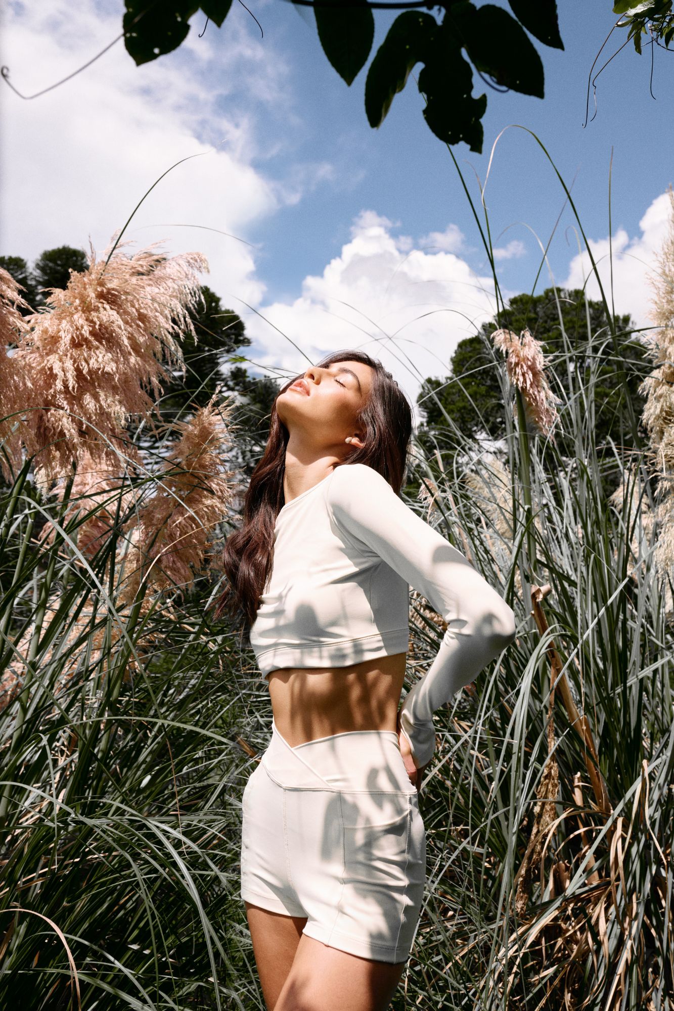 Mulher Yamoné de branco em meio à natureza, com gramíneas e céu azul.